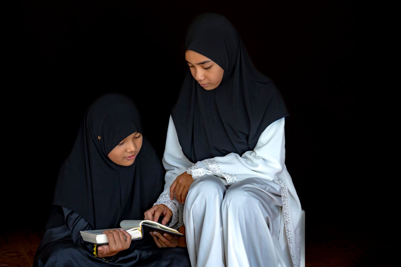 services-01 Two young women in traditional attire reading a holy book together, cultivating knowledge.